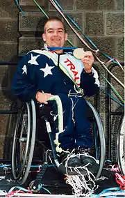 Homme en survêtement dans un fauteuil roulant, montrant une medaille devant lui.