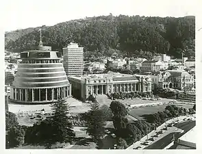 Photographie en noir et blanc d'un bâtiment de forme tronconique et de style moderne placé à gauche d'un bâtiment officiel de style néoclassique.