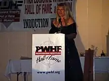 Une femme blonde en robe noire qui fait un discours derrière un pupitre.