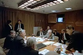 Des hommes en costume assis autour d'une table ovale discutent. Des papiers sont sur la table. Au fond, une télévision des années 1980 est allumée.