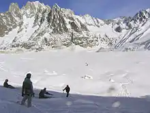 Skieur et snowboardeurs dominant une vaste étendue recouverte de neige au pied de sommets abrupts.