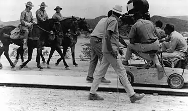 Photographie montrant l'équipe de tournage, composée de cinq hommes, maniant une caméra montée sur un véhicule léger. Ils filment trois acteurs en costume de soldats américains, montés sur des chevaux.