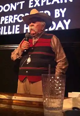 Un homme avec une grande moustache grise, un chapeau de cowboy et un pull rayé horizontalement rouge et noir. Il tient un micro dans sa main droite.