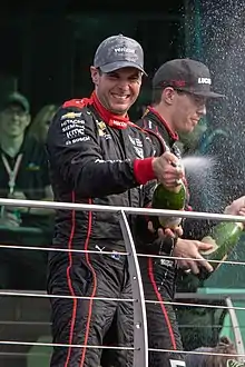 Homme, debout de face, ouvrant une bouteille de champagne pour célébrer une victoire. Derrière lui, un autre piolote fiat de même.