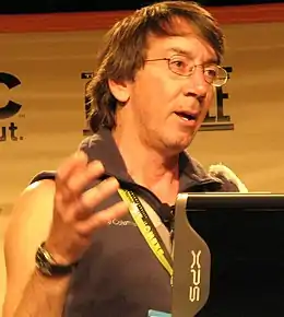 Will Wright parle dans un micro lors d'une conférence. Un écran de prompteur est visible à sa droite.