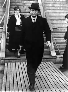 Un homme marche d'un pas décidé, en manteau noir, cravate et coiffé d'un chapeau