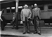 Photographie noir et blanc de deux hommes debout sur un quai avec un train derrière.