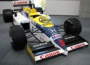 Photographie en couleur d'une Formule 1 bleu foncé, blanche et jaune, vue de trois-quarts droite, exposée dans un musée.