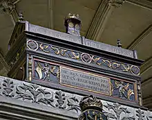 Photo d'un coffre oblong de couleur sombre décoré de motifs dorés, avec des inscriptions en latin. Il est surmonté d'une couronne.