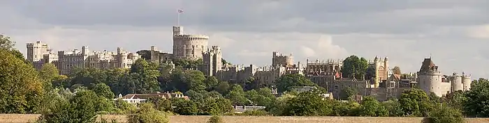 Panorama d'un château en pierre grise avec des arbres au premier plan et une tour imposante est visible au centre de l'image.
