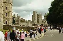 Photographie d'un château en pierre s'étendant à gauche de l'image. De nombreuses personnes marchent sur une route au centre.