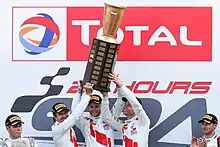 Photographie de trois hommes debouts et souriants, levant à eux trois un énorme trophée, sur un podium.