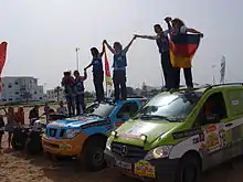 Déux véhicules automobiles vus de côté. Les pilotes et copilotes de ceux-ci sont debout sur leur toit, avec leurs drapeaux nationaux.