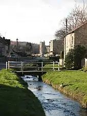 Photo d'une rivière bordée par des talus enherbés. Sur la droite de la rivière se trouve une maison en pierre envahie par la végétation, avec un toit en tuiles oranges et deux fenêtres blanches. La maison est accessible par un pont en béton avec des barrières blanches passant au-dessus de la rivière. Sur la gauche de la rivière se trouve une route goudronnée qui mène, au loin, à un château en pierres avec deux tours carrées.