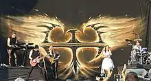 Photo en couleur d'un groupe de musique sur scène, à l'arrière-plan un logo argenté sous forme d'un oiseau de feu