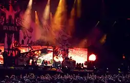 Photo en couleur montrant un groupe de musique sur scène, à l'avant plan des spectateurs lèvent les mains