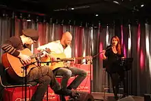 Photo en couleur d'un groupe jouant de la musique dans une pièce, deux guitaristes sont assis à gauche, une chanteuse aux cheveux bruns est debout derrière un pupitre à droite