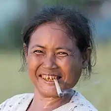 Portrait d'une femme serrant une cigarette roulée à la main entre ses dents, au coucher du soleil dans la campagne de Don Det, Laos. La couleur rouge de ses dents est due à la mastication régulière de paan. Septembre 2017.