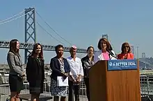 Photographie couleur des représentantes du Congrès pendant un discours, devant un pont de San Francisco.