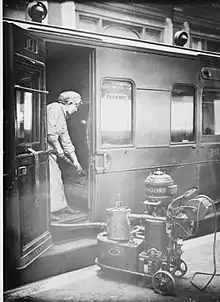 Photo ancienne : Une femme en robe et bonnet, debout dans une voiture de chemin de fer à la portière ouverte, nettoie une banquette à l'aide d'un très gros aspirateur à roulettes resté posé sur le quai.