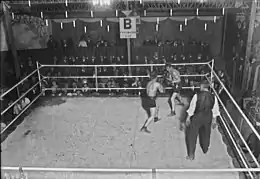 Photographie en noir et blanc de deux boxeurs s'affrontant sur un ring devant quelques rangées de spectateurs habillés en costumes.