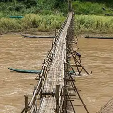 Vue de face d'une passerelle en bois traversant la rivière Nam Khan, avec un ouvrier travaillant à la consolidation de cette structure et se tenant témérairement debout sur un pilier extérieur incliné, à Luang Prabang. Juin 2018.