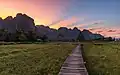 Passerelle en bois élevée sur pilotis, menant vers une hutte au toit de paille, en face des montagnes karstiques, au coucher du soleil avec un ciel coloré, à Vieng Tara Villa, Vang Vieng, durant la mousson.