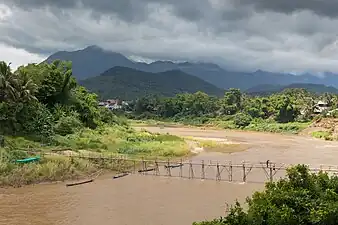 Paysage avec une passerelle en bois traversant la rivière Nam Khan. Deux ouvriers travaillent à sa consolidation, portant une lourde poutre sous un ciel orageux durant la mousson, à Luang Prabang. Juin 2018.