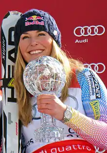 Femme blonde, souriante, portant un bonnet, une paire de ski appuyée sur son épaule droite et un trophée dans la main gauche