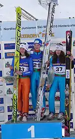 3 jeunes filles côtes-à-côtes sur la plus haute marche d'un podium de sport, en combinaison de saut à ski, tenant chacune leur skis de saut verticalement, avec un serre-tête aux couleurs de leur sponsor