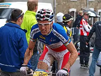  Homme sur un VTT en tenue bleu-blanc-rouge maculée de boue avec des spectateurs en arrière-plan.