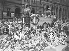 Photographie d'un bloc de granit entouré par deux statues de soldats. Le monument est recouvert de gerbes de fleurs et une foule nombreuse, majoritairement composée de femmes, est présente.
