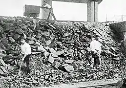 Photo en noir et blanc montrant des hommes travaillant sur une pente de pierre, au dessus un homme pousse un wagonnet.