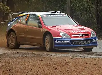 Photo de la Citroën Xsara WRC de Sébastien Loeb, à la livrée rouge, blanche et bleue, vue de trois-quart en dérapage dans un virage boueux.