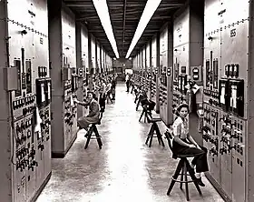 Un long couloir dans lesquelles des femmes assises sur des tabourets hauts surveillent des consoles avec des cadrans et des interrupteurs.