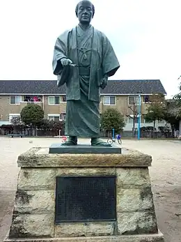 Photo couleur d'une statue de bronze sur un socle en pierre figurant un homme debout en habits traditionnels japonais.