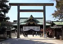Photo couleur d'un torii (au premier plan) et du bâtiment réservé au culte dans l'enceinte d'un sanctuaire shintō. En arrière-plan, un ciel bleu.