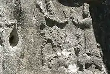 photo d'un mur sculpté, on voit deux hommes face à face tenant chacun une coupe, aux pieds du deuxième, un chien