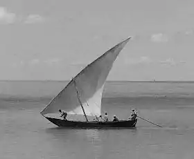 Photographie en noir et blanc d'un navire en mer avec une voile tendue, de forme triangulaire, et plusieurs occupants.
