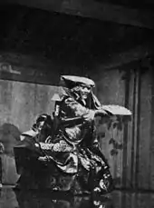 Acteur en tenue traditionnelle, masque féroce, bras en avant tenant un éventail. Noir et blanc.