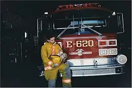 A firefighter holds a baby, in front of a 1990 American LaFrance Century 2000 pumper.