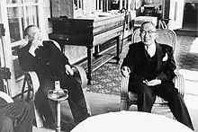 Photo noir et blanc de deux hommes en costume sombre, assis devant une table ronde.