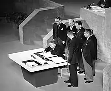 Photo noir et blanc d'un homme en costume sombre, assis devant une table blanche, un stylo à la main. Six hommes en costume sombres se tiennent debout derrière lui.