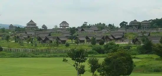 Photo couleur d'un ensemble de constructions en bois et toit de chaume, dans une plaine arborée et verdoyante, sur fond de ciel nuageux.