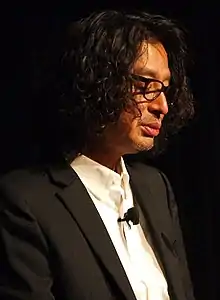 Portrait de Yoshio Sakamoto. Habillé d'une veste de costume noire et d'une chemise blanche, il est pris légèrement sur son profil droit.