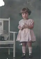 Photographie colorisée. Janet Rosenberg, alors enfant en bas âge, pose debout en robe courte lors d’une séance de photographie. À gauche, un aquarium est posé sur une chaise. Le fond est de couleur terne.