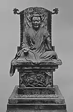 Vue de face d'une statue assise les jambes croisées placé sur un trône. Son bras droit repose sur sa jambe, le bras gauche est légèrement surélevé et plié.
