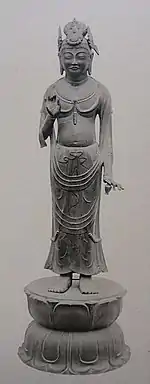 Vue de face d'une statue sur un piédestal avec la main gauche levée à hauteur de poitrine. La main gauche porte un petit objet en forme de vase ou de pot.