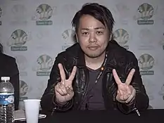 Homme japonais aux cheveux courts, en train de faire un signe des mains.