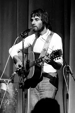 Photo noir et blanc d'un homme barbu aux cheveux mi-longs jouant de la guitare.
